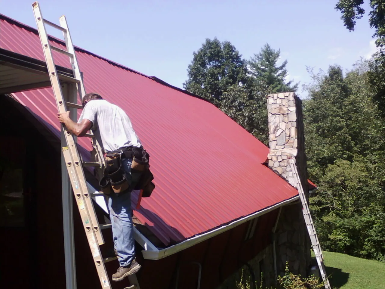 Roofer on a ladder beside a red metal roof and stone chimney — Soffit & Fascia Repair by Roofing Brothers in Wyola